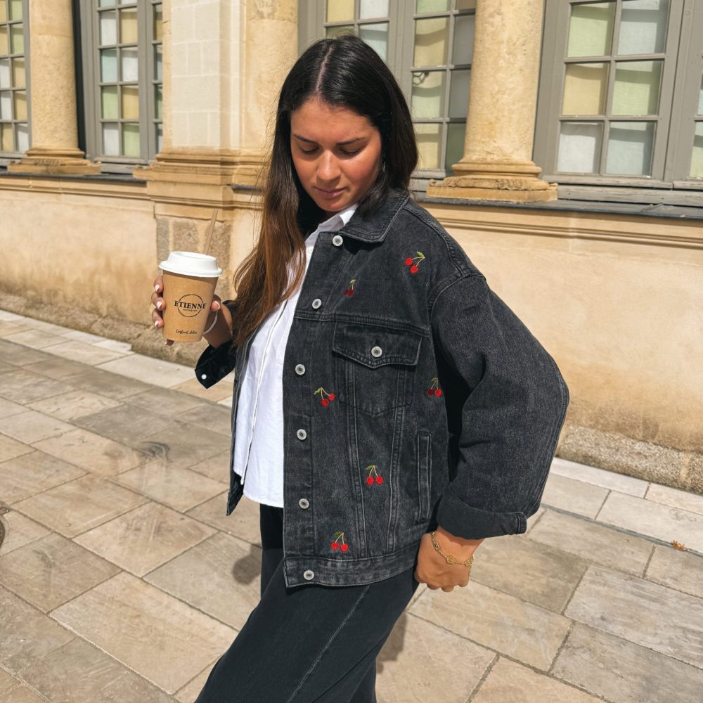 Femme portant une veste en jean avec de la broderie motifs cerise, avec un gobelet et une paille