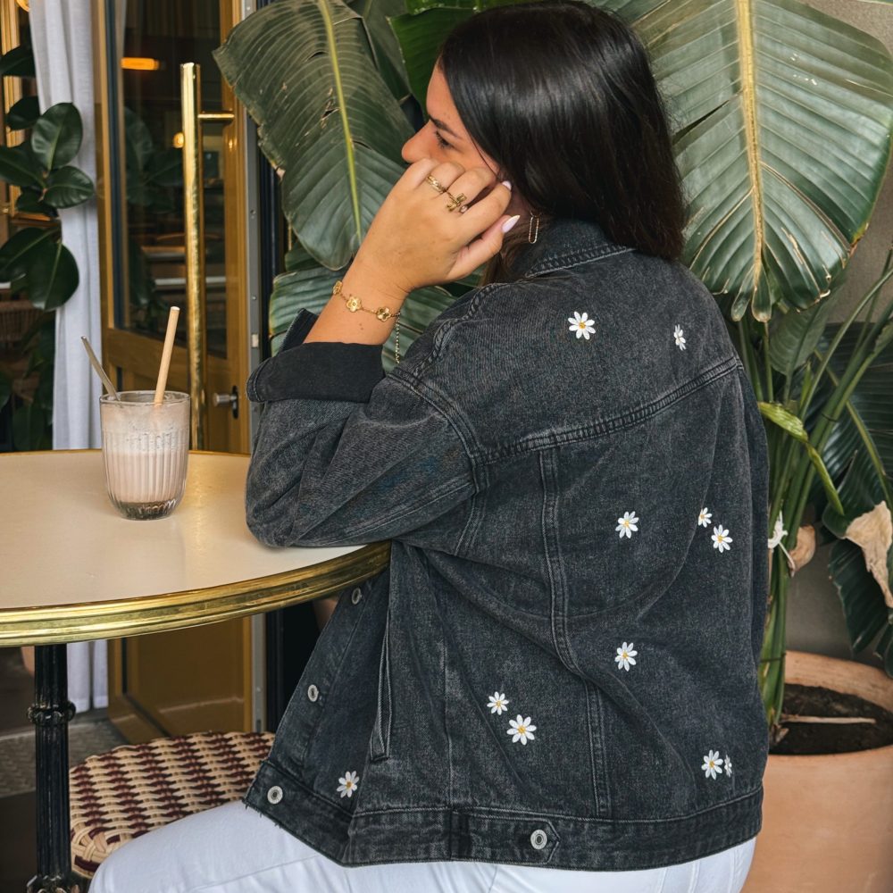 Femme portant une veste en jean avec de la broderie motifs marguerites, sur un tabouret dans un bar