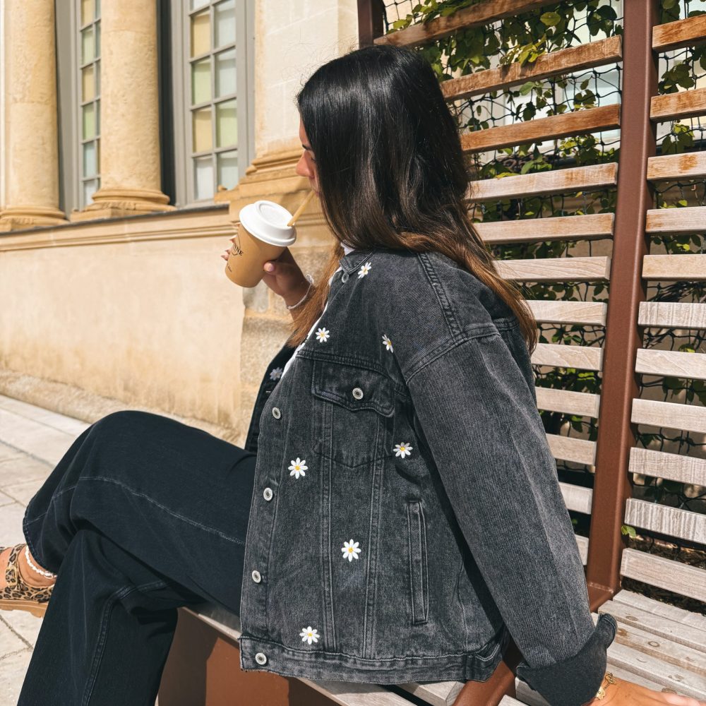 Femme portant une veste en jean avec de la broderie motifs marguerites, sur un banc en extérieur